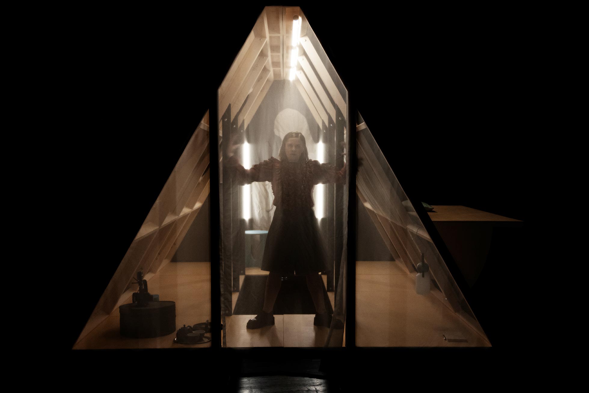 Person steht in einem beleuchteten, dreieckigen Raum mit Holzstruktur und transparenten Wänden