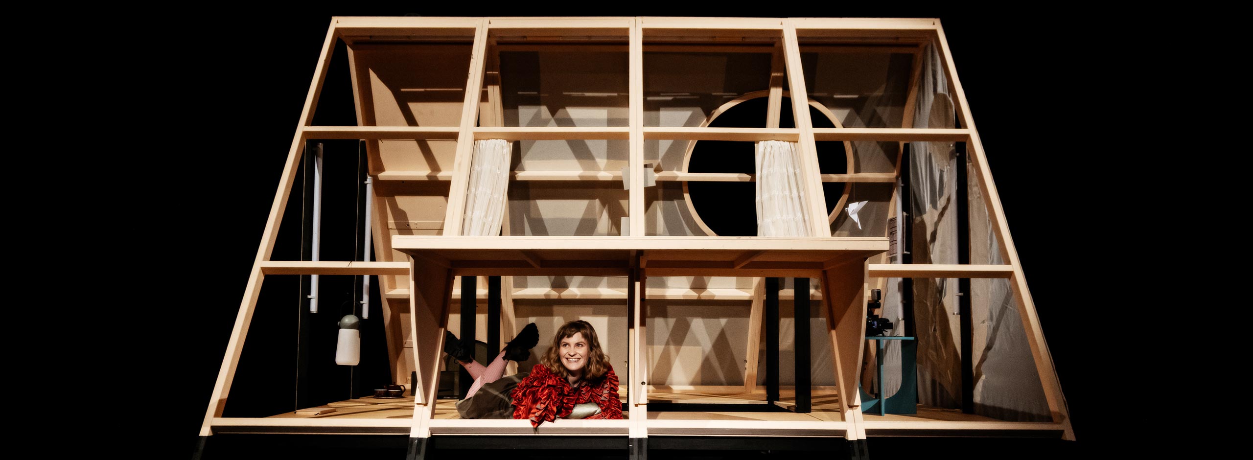 Frau in rotem Oberteil liegt lächelnd in einem offenen, zeltähnlichen Holzrahmen auf einer Theaterbühne