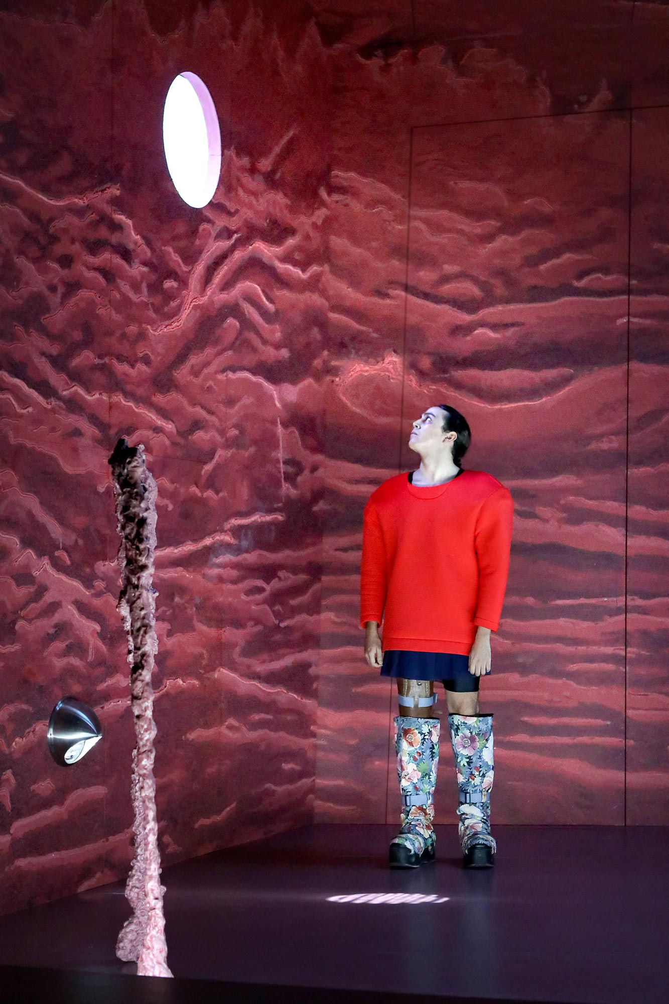 Person in rotem Pullover und gemusterten Stiefeln steht auf Theaterbühne und blickt nach oben<br />
