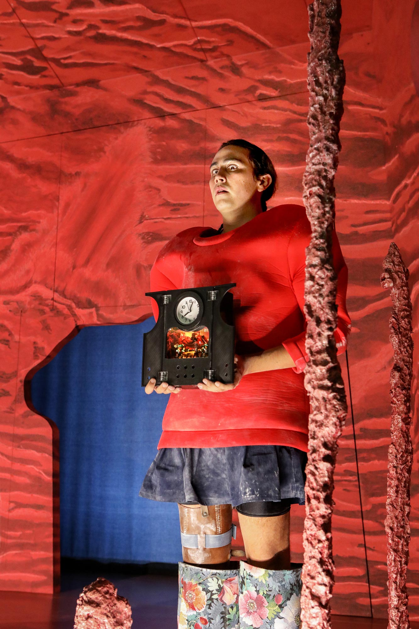 Person mit rotem Kostüm hält schwarze Uhr mit Miniatur-Szene vor surreal rotem Bühnenhintergrund