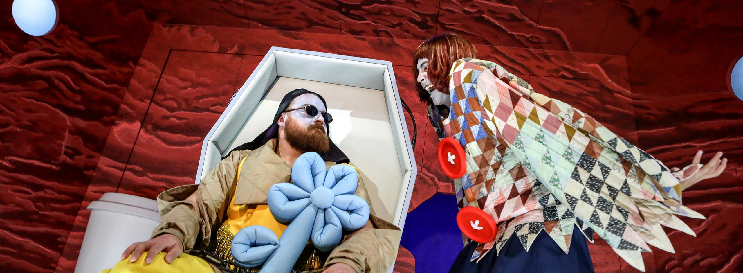 Ein Schauspieler liegt in einer futuristischen Liege mit Sonnenbrille und Plüschblume, während eine andere Figur mit buntem Kostüm sich über ihn beugt.
